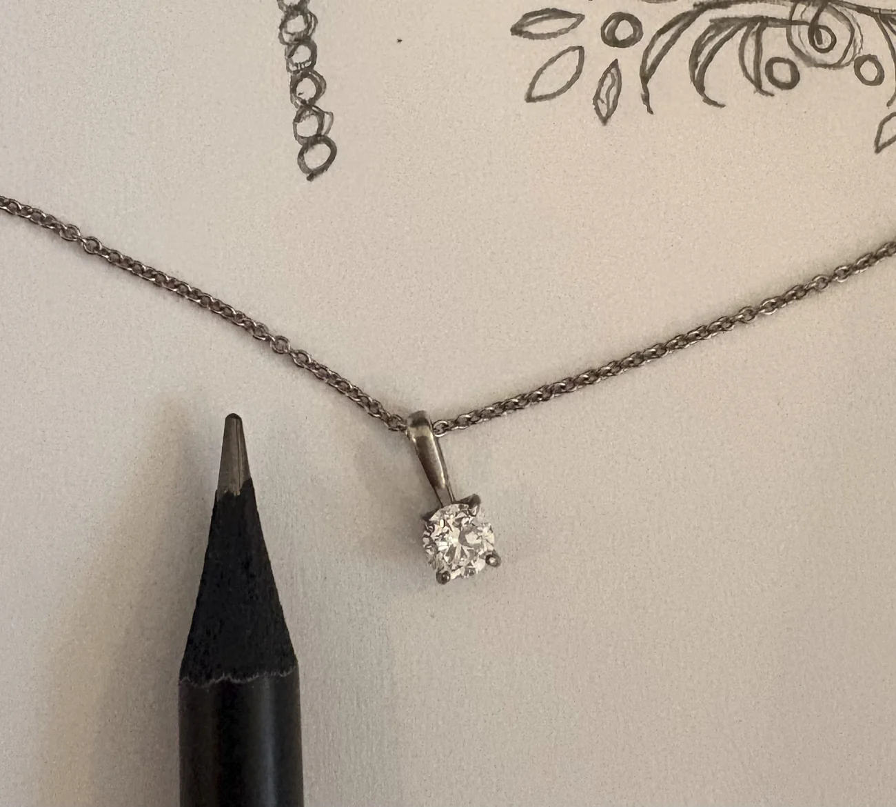 A diamond solitaire pendant on a fine chain lying on a sketchbook page beside the sharpened tip of a graphite pencil