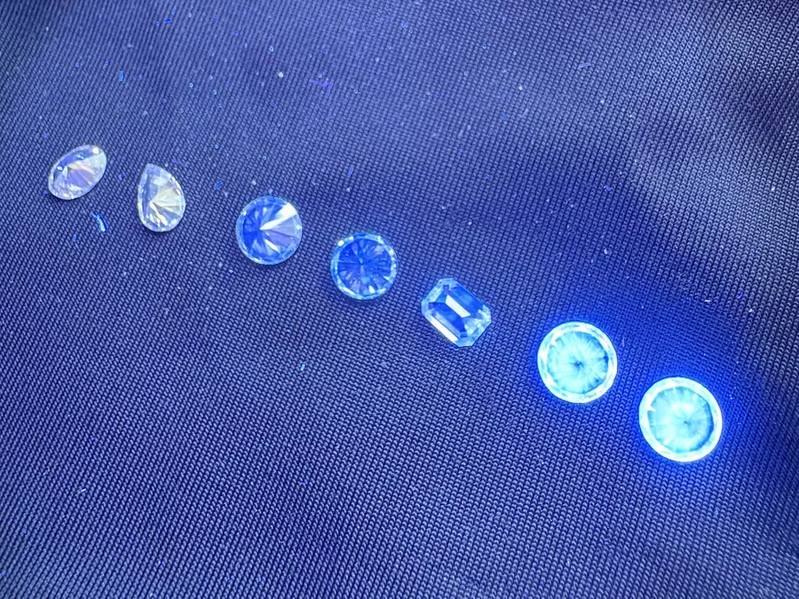 Seven colourless diamonds laid in a row on dark fabric under an ultraviolet lamp. The two at the right glow a bright blue-white; the glow weakens along the row until the two at the left barely react.